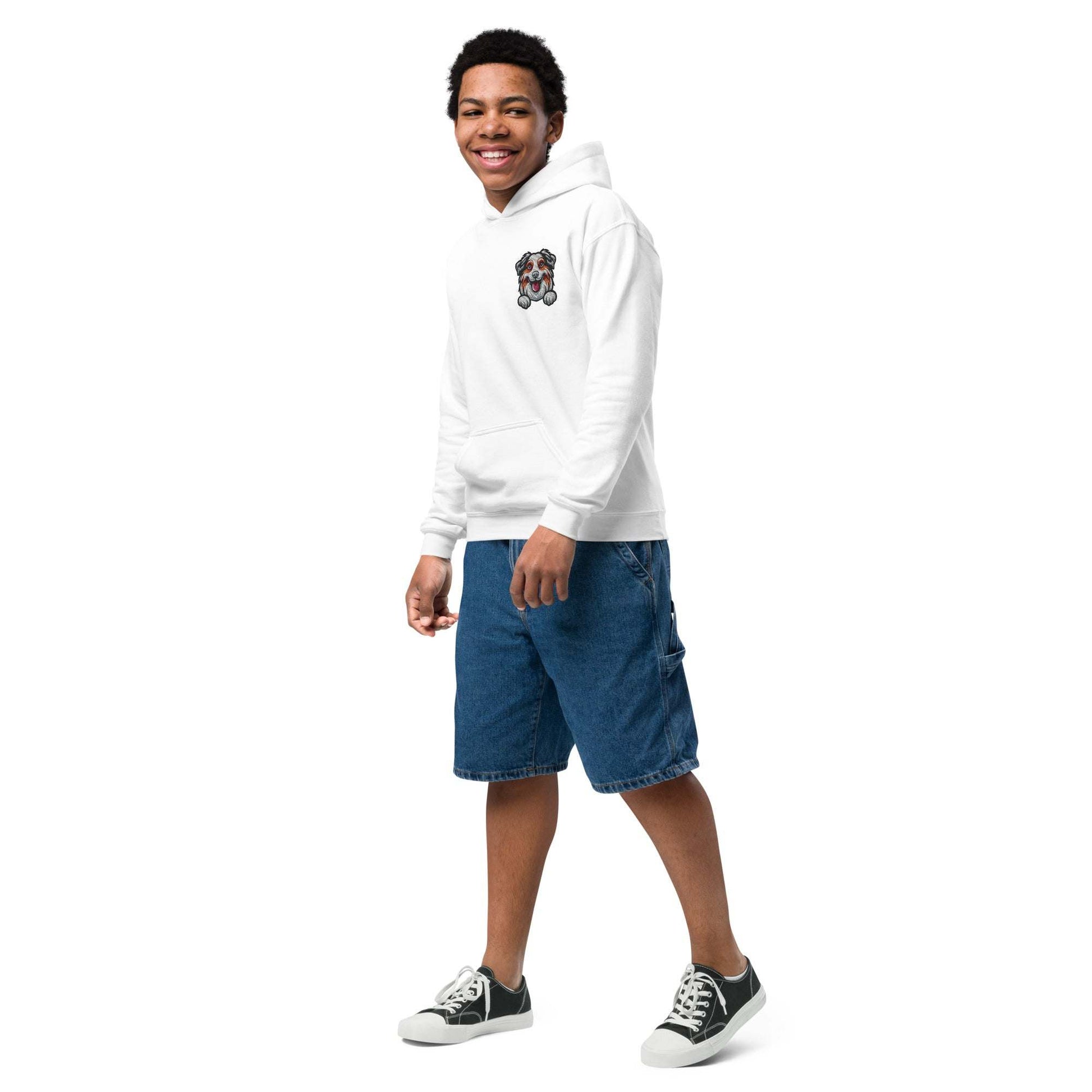 Young man wearing white premium youth hoodie with embroidered Australian Shepherd dog design, paired with denim shorts and sneakers